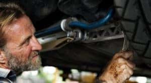 A bearded man is working on a lifted vehicle.