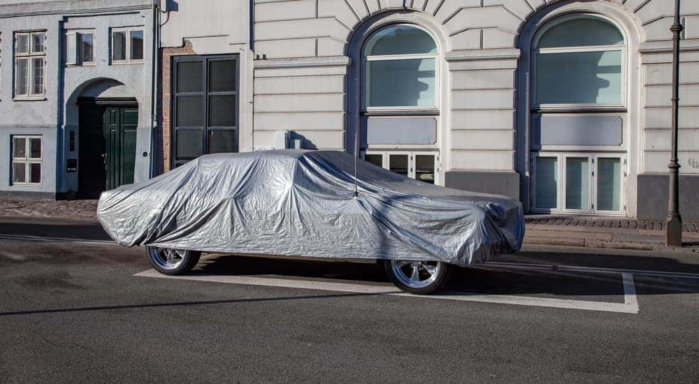 A sedan under a gray car cover in front of a white building