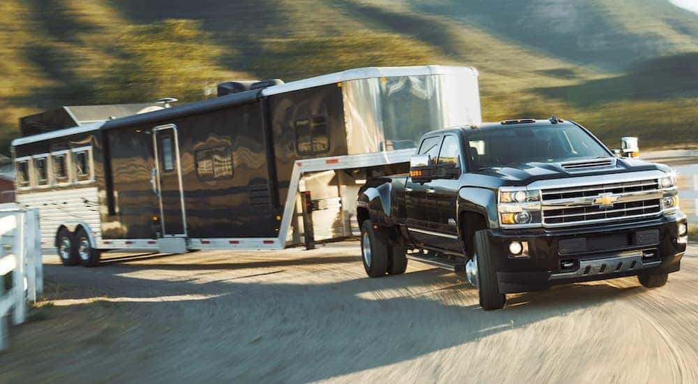 A black 2019 Chevy 3500 is towing an enclosed goose neck trailer on a dirt, farm drive way.