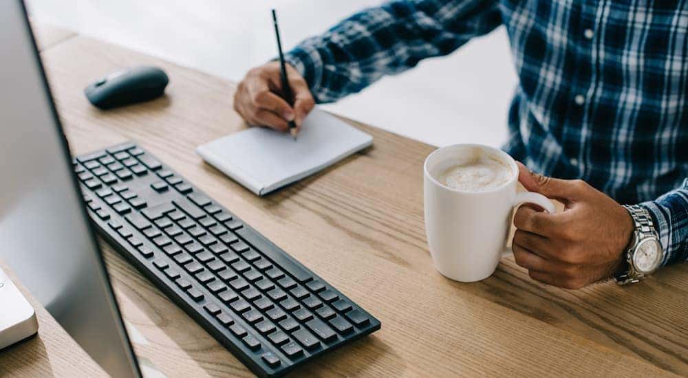 A man's hands can be seen writing on a note pad with a computer in front of him and a coffee in his other hand.