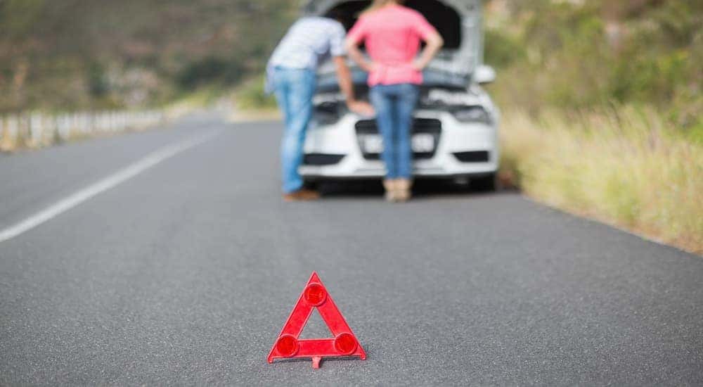 An orange warning marker is set up with a blurred couple and their broken car behind it.