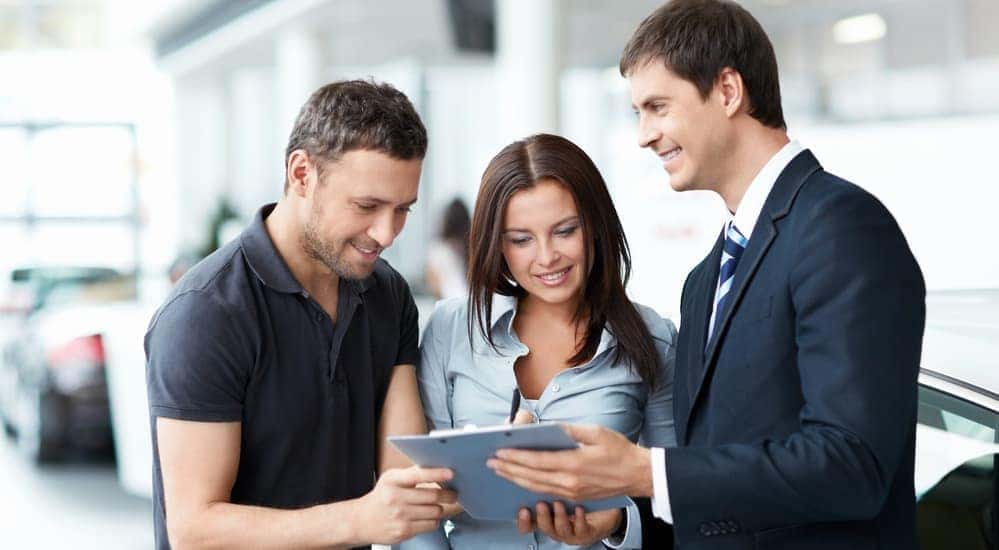 After searching, 'Used Cars in Columbus, Ohio', a smiling couple is at a used car dealership signing sales papers with a car salesman.