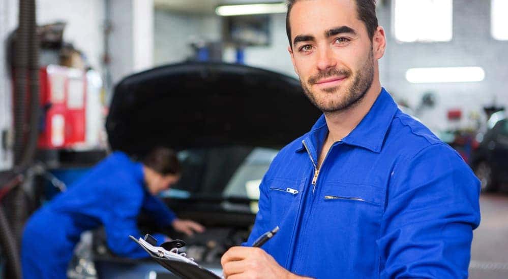 A smiling mechanic, who is working at a best rated Chevy dealer near me, is standing in a garage with a blurred car behind him.
