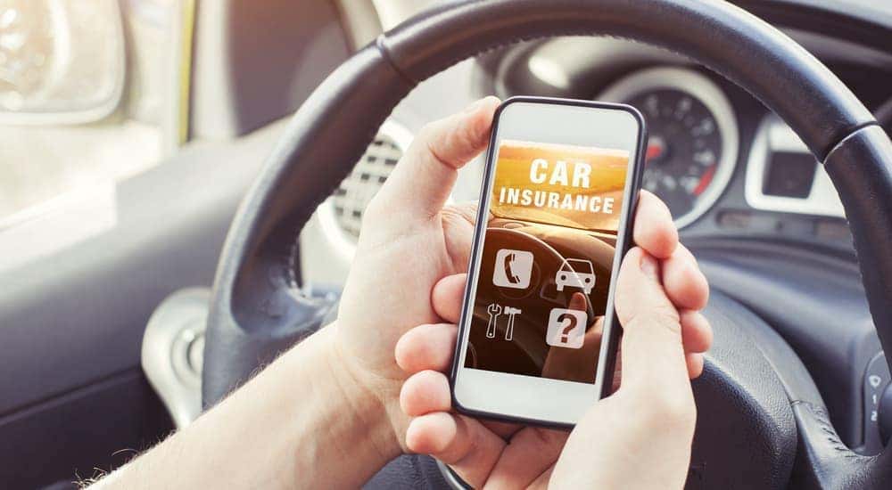 A set of hands in front of a car steering wheel is holding up a phone that says car insurance on it.