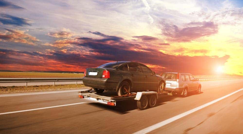 A small silver SUV is towing a used car on a trailer toward a sunset near Cincinnati, OH.