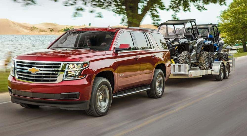 A red 2020 Chevy Tahoe is driving on a tree lined road while towing two side by sides on a large trailer.
