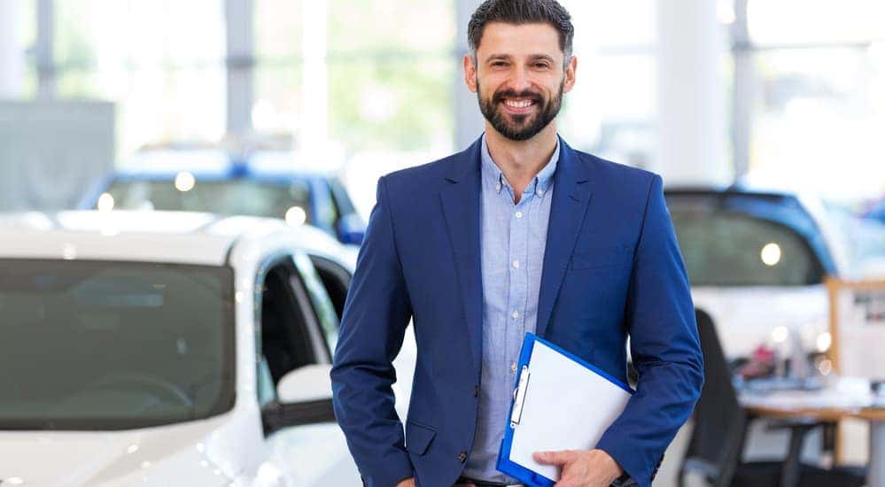A smiling car salesman, who is similar to someone that you'll see while shopping for used cars for sale in Ohio, is standing in a car showroom.