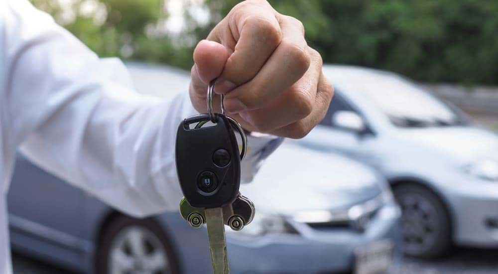 A hand is holding a set of used car keys with a blurred car in the background. 