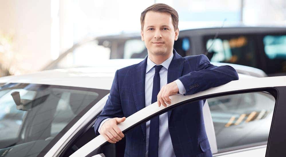 A smiling car salesman is standing behind the door of a used car.