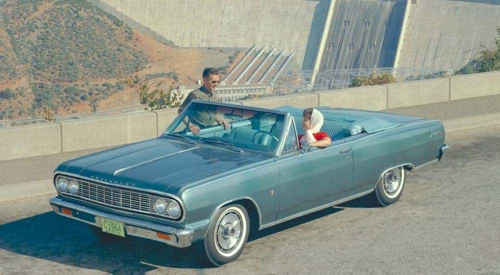 A blue 1964 Chevy Malibu convertible is parked in front of a dam.