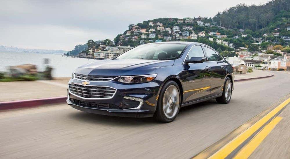 A popular used car for sale, a blue 2018 Chevy Malibu, is driving on a coastal road.