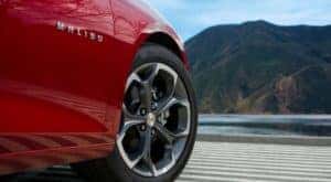 A closeup shows the tire and side badging on a red 2018 used Chevy Malibu that is parked in front of a river and mountain.