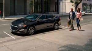 A couple is walking past a black 2021 Chevy Malibu that is parked on a street.