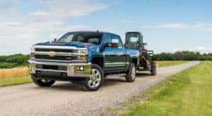 A blue used diesel truck, a 2018 Chevy Silverado 2500HD, is towing construction equipment on a dirt road.