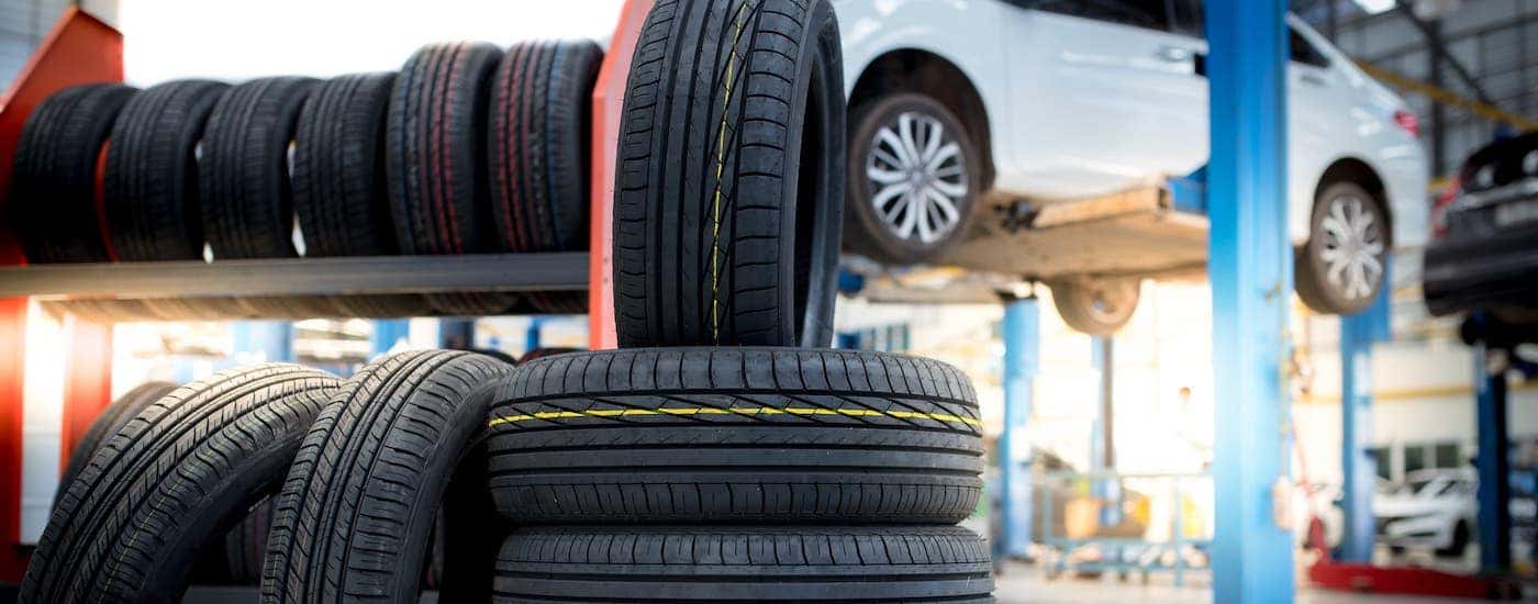 A pile of tires and tire rack are shown in a garage in front of a white car on a lift.