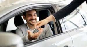 A salesperson is handing keys to a man inside a silver car.