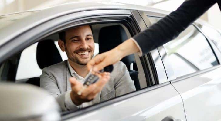 A salesperson is handing keys to a man inside a silver car.