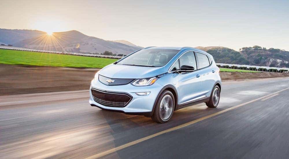 A light blue 2018 Chevy Bolt EV is driving down the highway with a sun cresting the mountains in the background.