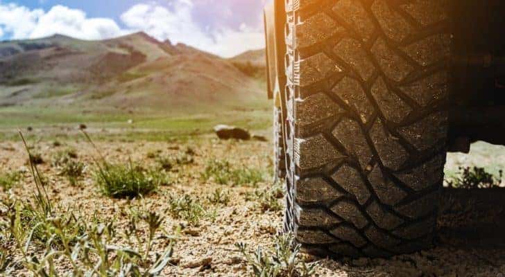 A closeup shows a tire on a vehicle overlooking a field and distant mountains.