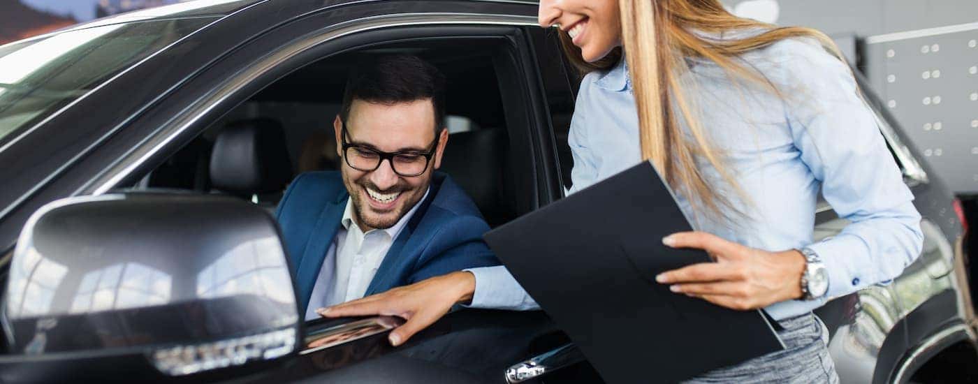 A saleswoman is showing a man features while he sits in a grey vehicle.