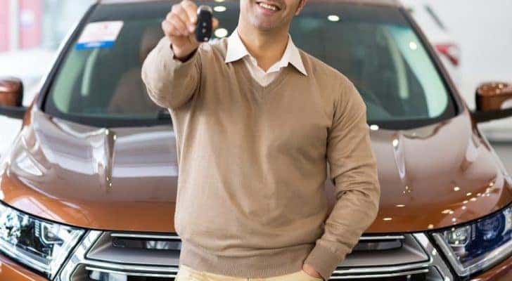 A man is leaning on an orange SUV holding out keys at a used Ford dealer.