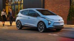 One of the most popular new cars in stock, a pale blue 2022 Chevy Bolt EV, is shown parked on a street in front of a brick building.