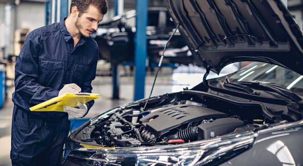 A mechanic is shown inspecting the engine of a grey sedan.