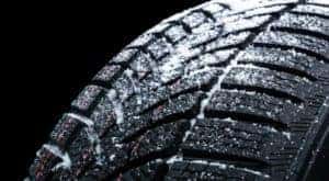 A close up of a winter tire with snow is shown on a dark background.