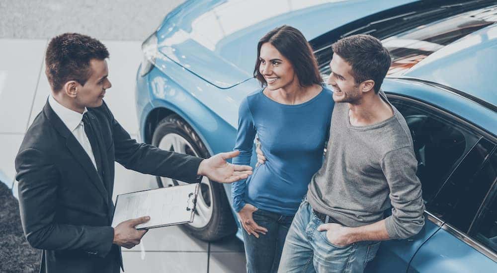 A salesman is holding a clipboard with paperwork while talking to a couple at the car dealership.