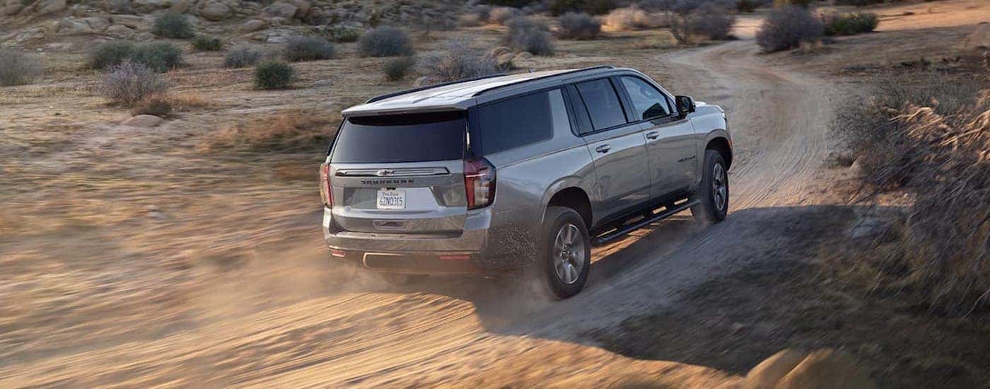 A popular used SUV for sale, a grey 2021 Chevy Suburban is shown from the rear driving on a dirt road.