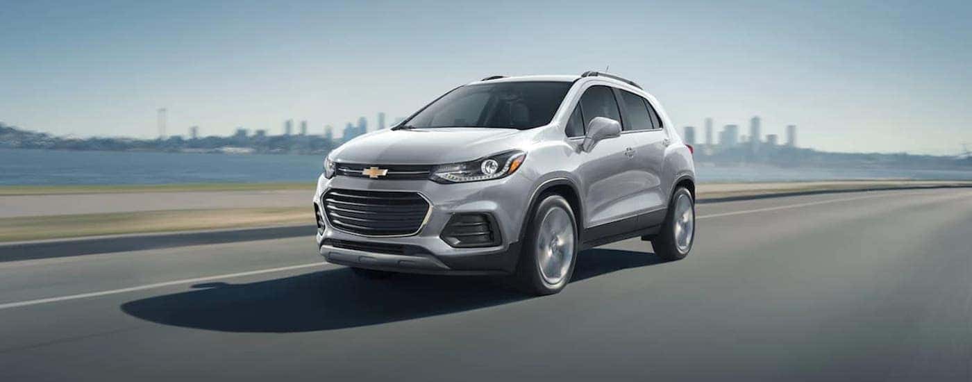 A silver 2021 Chevy Trax is shown from the front driving on an open highway past a city.