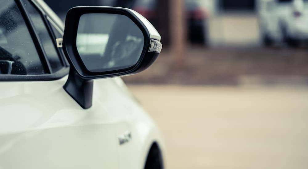 The side mirror on a white sedan is shown in a parking lot.