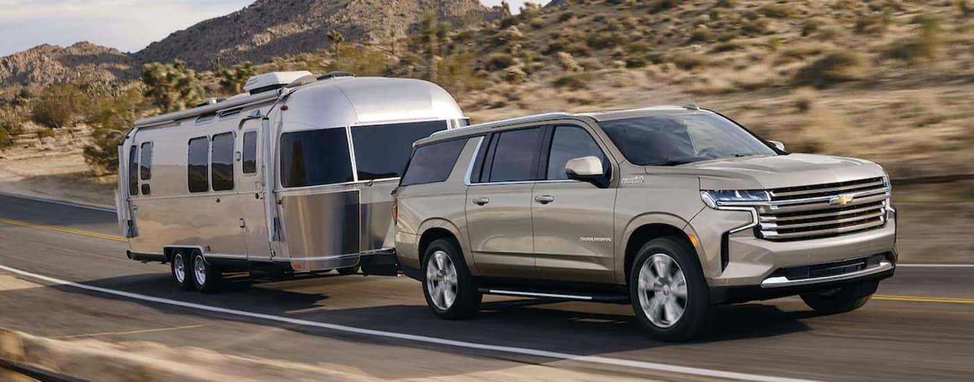 A tan 2023 Chevy Suburban is shown towing a camper trailer on an open road.