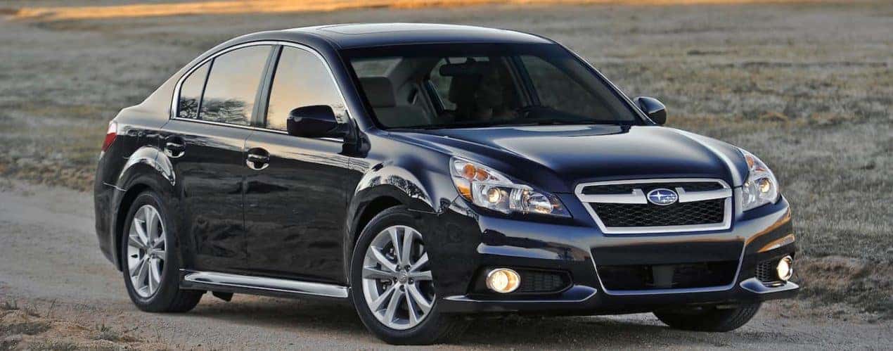 A black 2014 Subaru Legacy is shown driving on an open road next to a field.