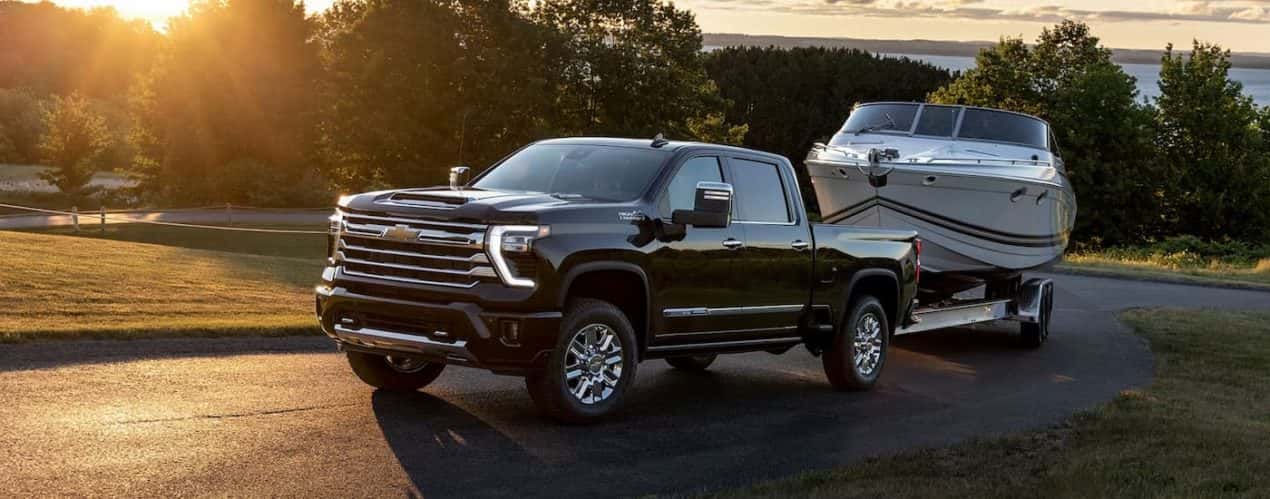 A black 2024 Chevy Silverado 2500 HD High Country is shown towing a white boat on a winding road.