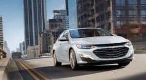 A popular Chevy car for sale, white 2023 Chevy Malibu, is shown driving on a city highway.