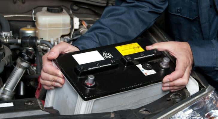A mechanic removing a used car battery from under the hood of a car.