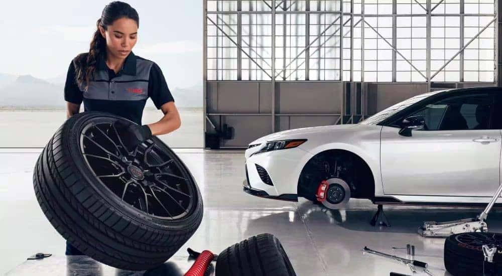 A person is shown changing the wheel on a 2023 Toyota Camry TRD at a Toyota service center in Cincinnati.