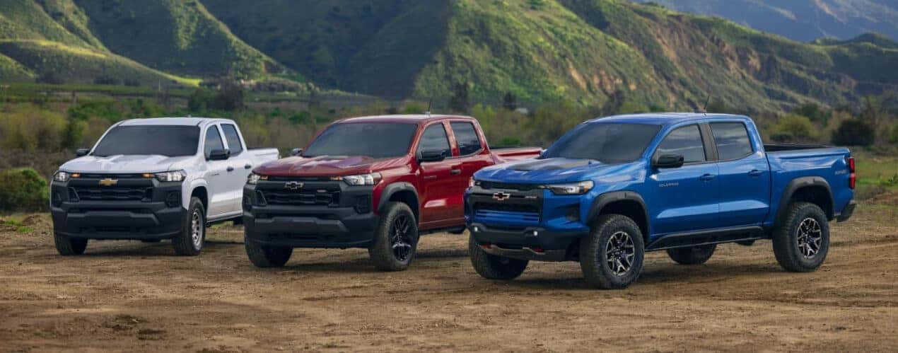 A white 2025 Chevy Colorado WT, a red Trailboss, and a blue ZR2 are shown on a clearing.