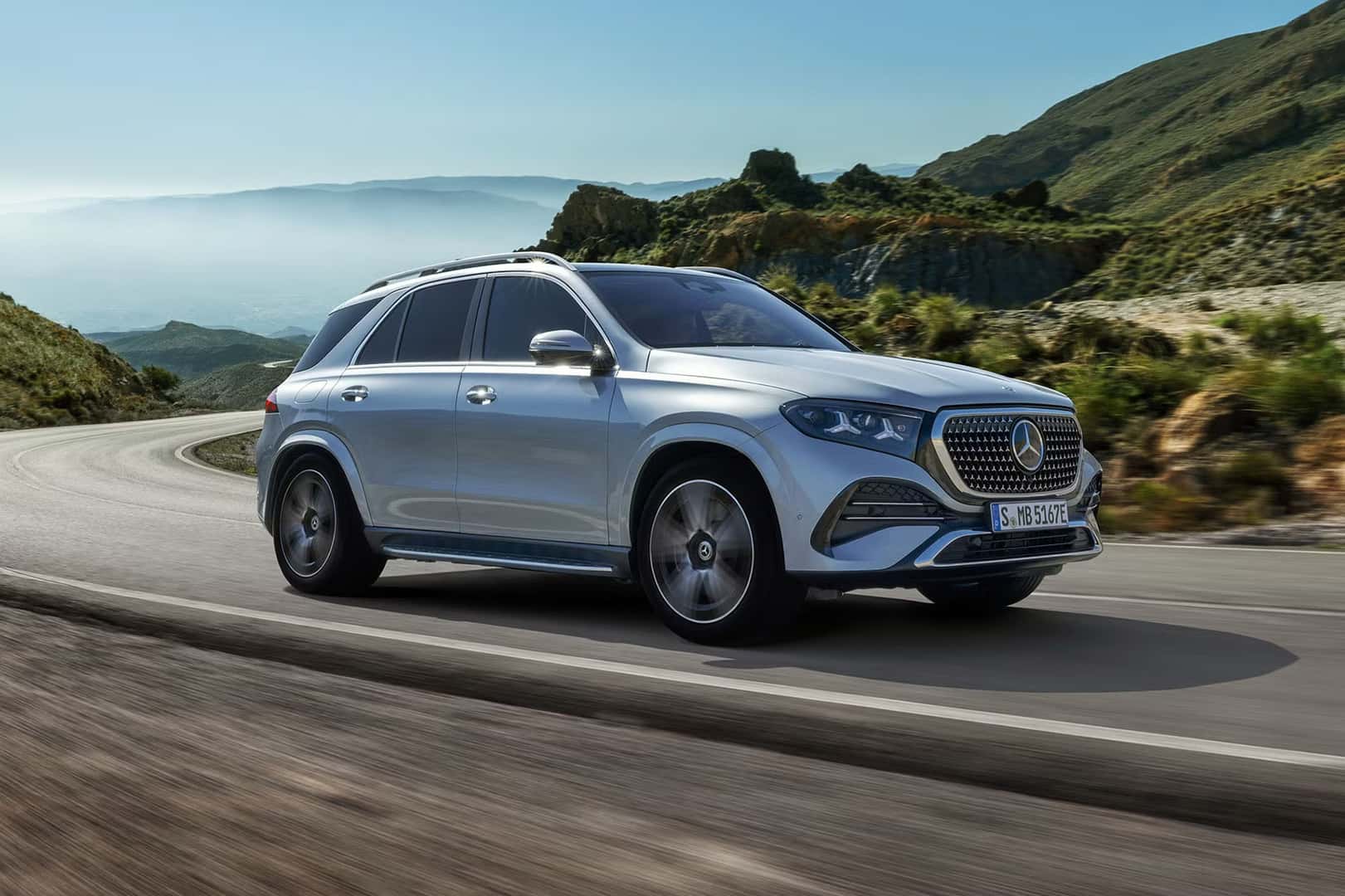 Silver 2027 Mercedes-Benz GLE SUV driving on a winding mountain road, front three-quarter view