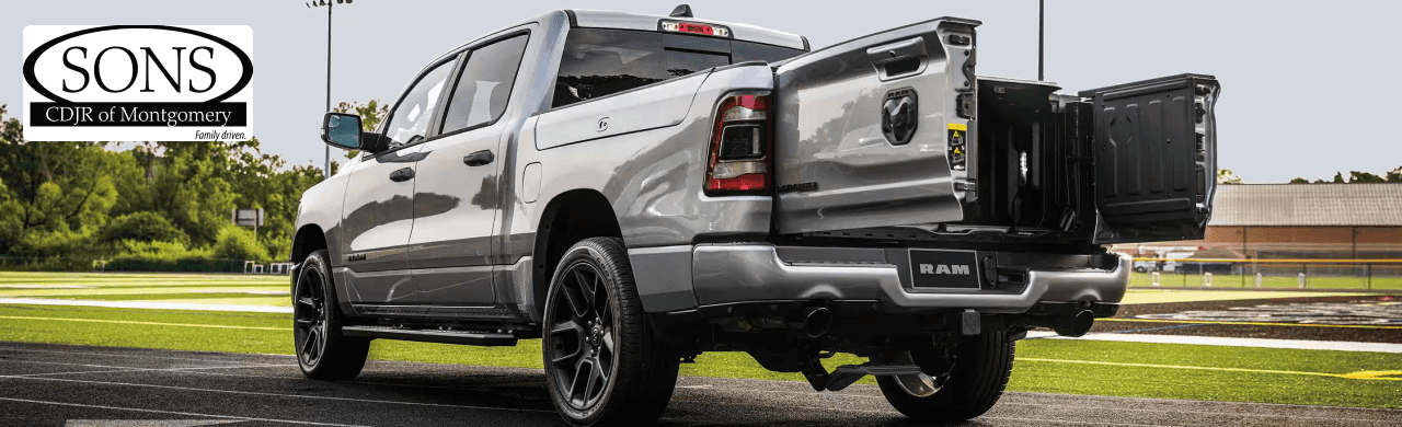 A gray Ram 1500 pickup truck is parked in a parking lot with its tailgate down, revealing a multi-function tailgate with the Sons CDJR of Montgomery logo in the top left corner.