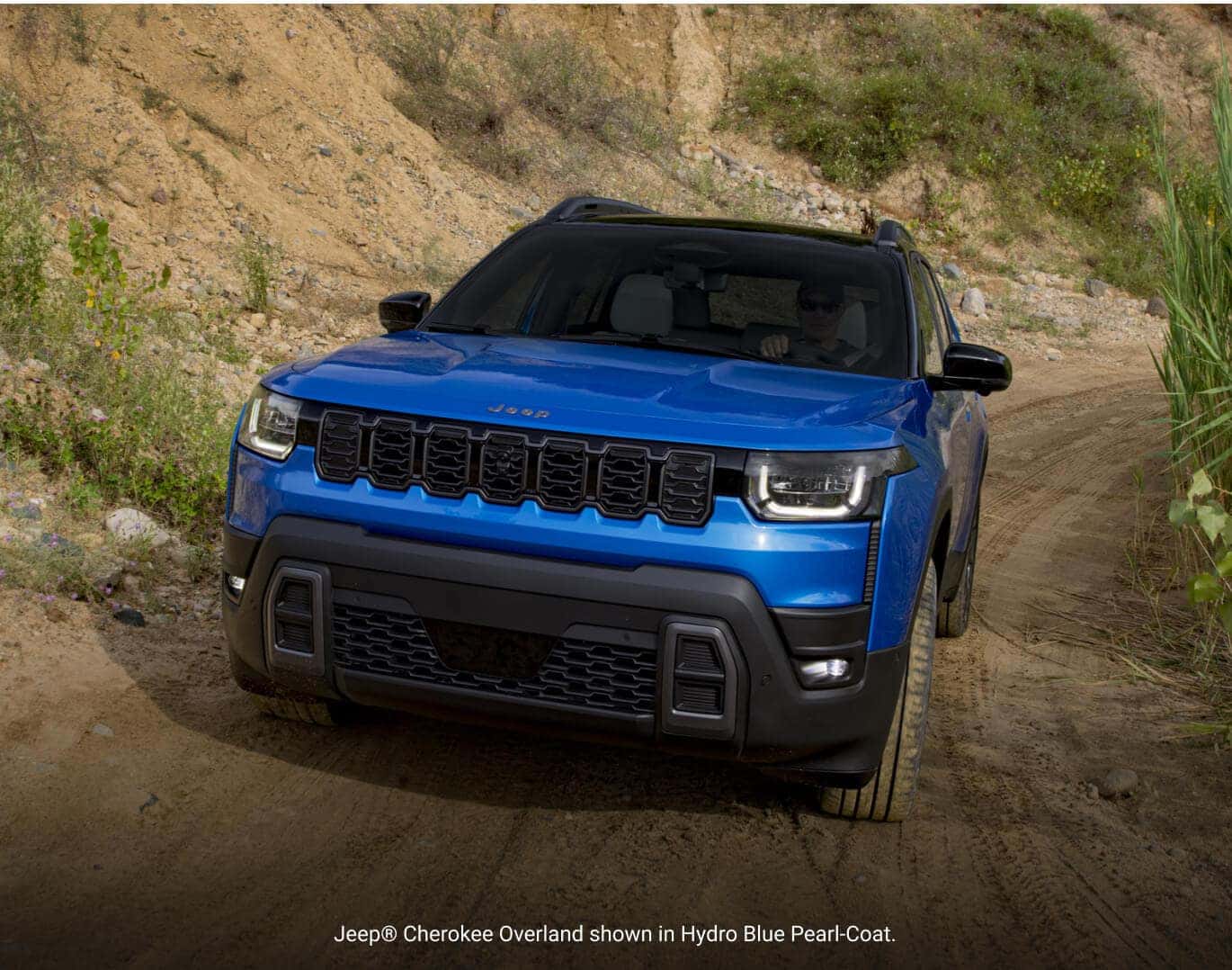 Front view of a 2026 Jeep Cherokee Overland in Hydro Blue Pearl-Coat driving along a dirt off-road path.