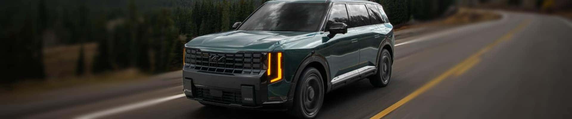 Green Kia SUV driving on a winding two-lane road surrounded by trees.